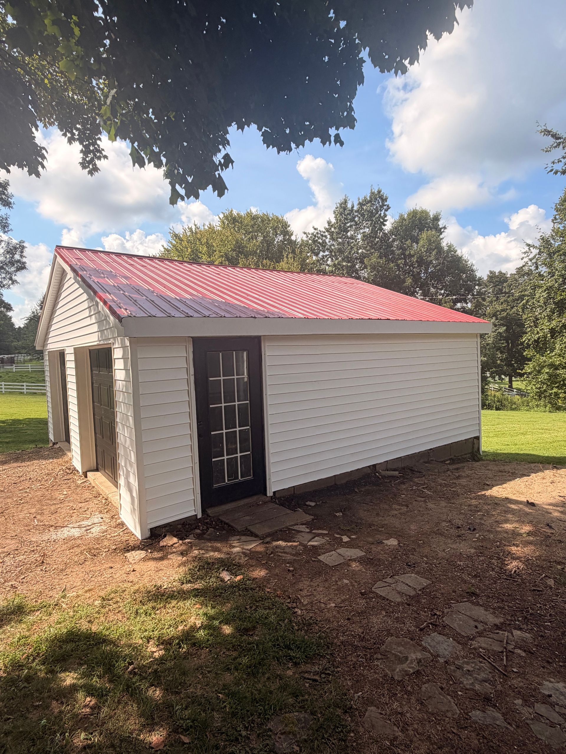 Small white brick outbuilding with red metal roof, black door, and trees overhead on a dirt lot.
