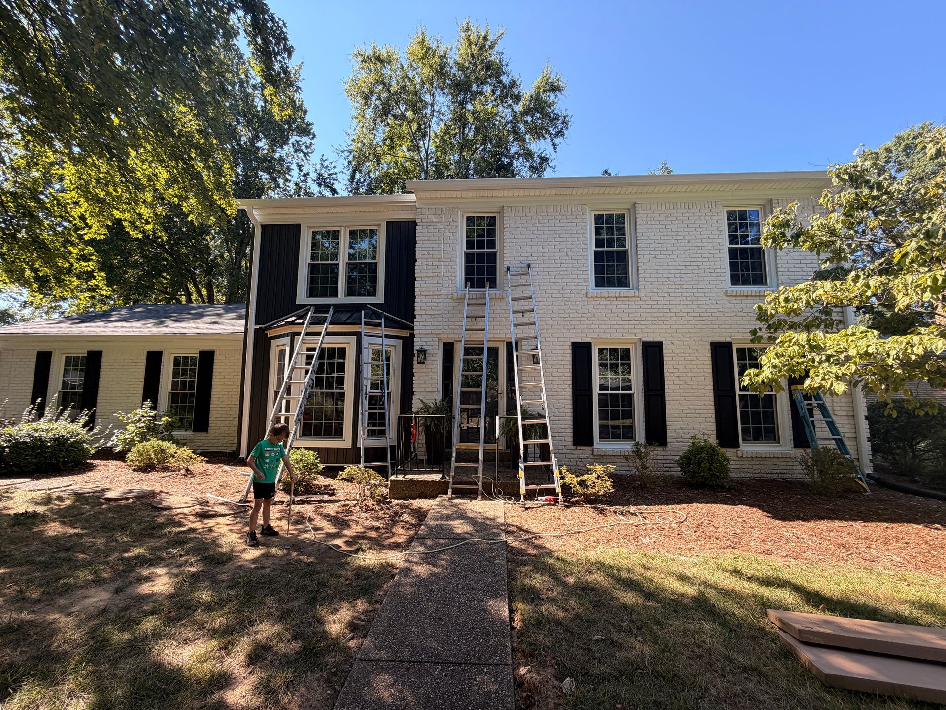 Two-story white house with black shutters, front steps, and a child standing on the walkway in a sunny yard