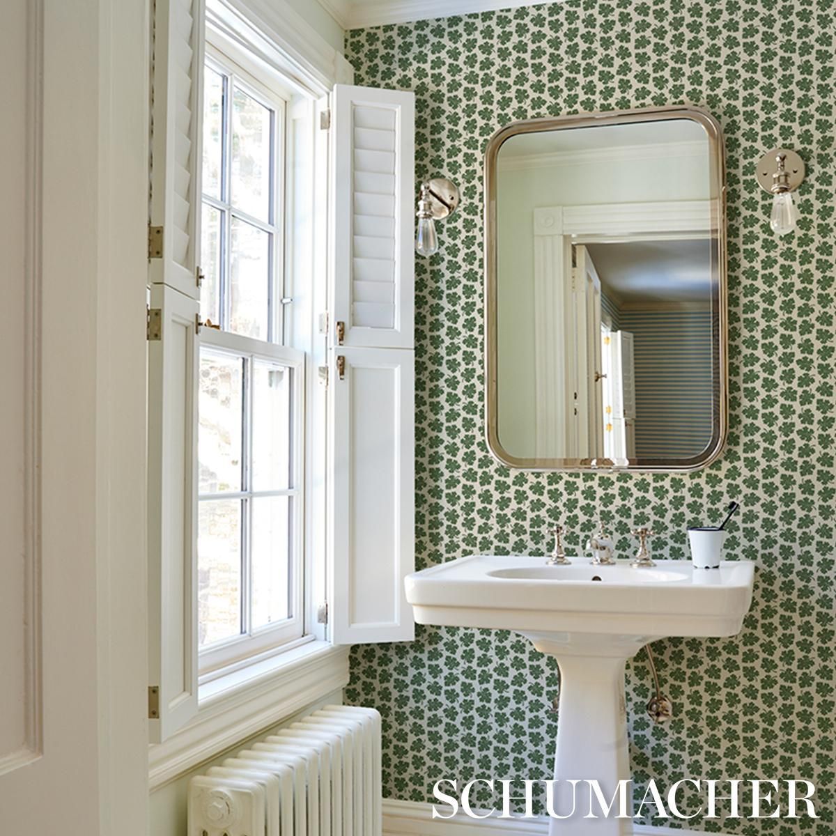 Bathroom with patterned green wallpaper, white pedestal sink, mirror, and window with shutters.