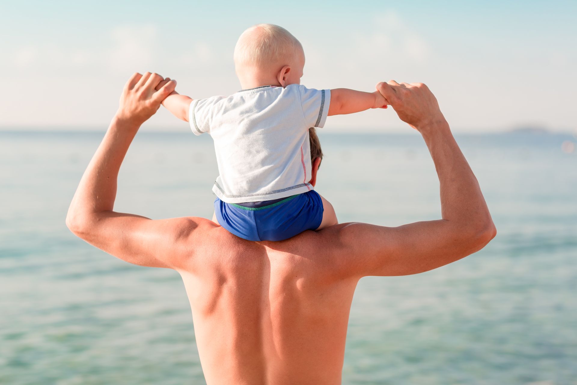 Father carrying baby on his shoulders at the beach, blue sea and sky in the background.