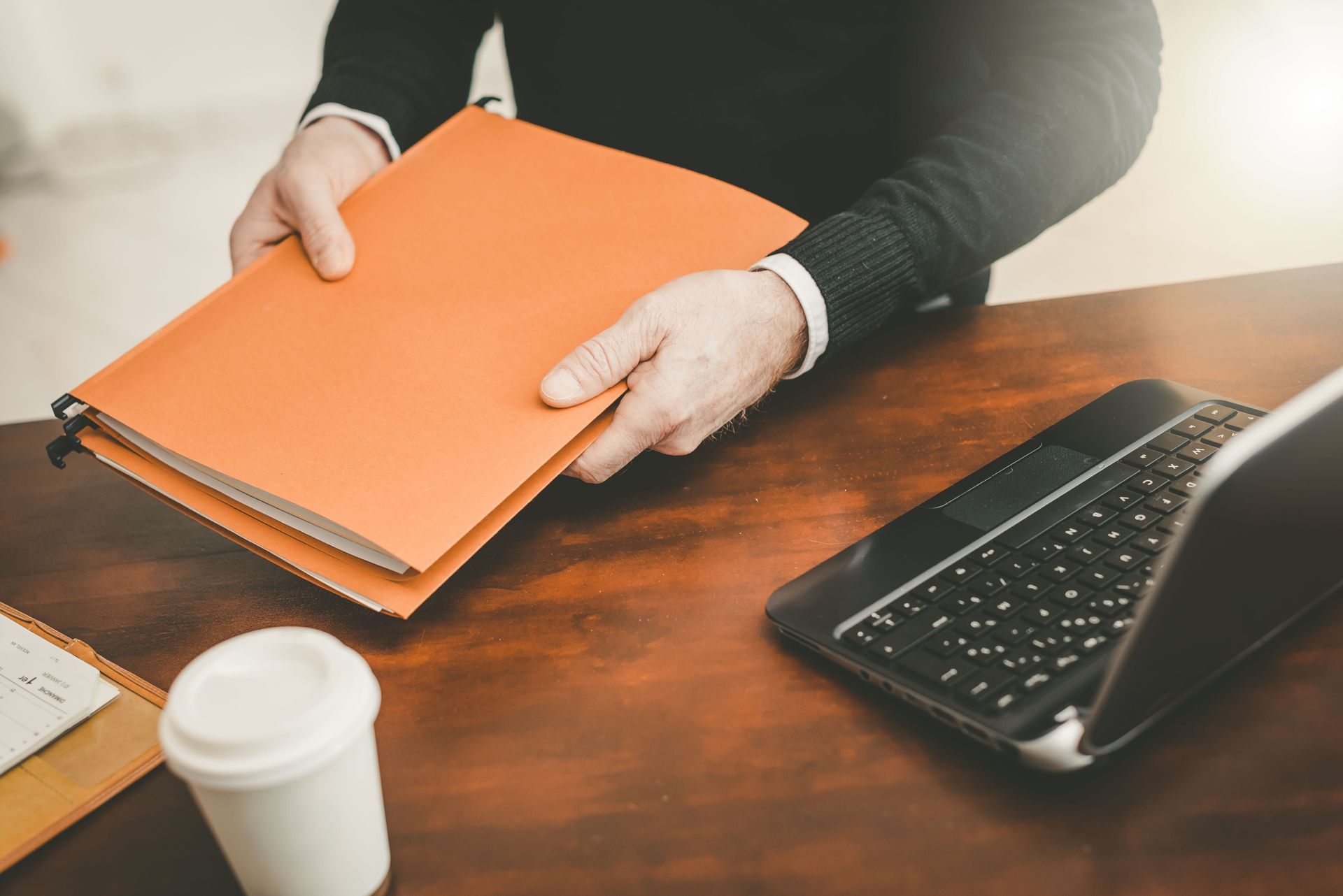 Hands holding an orange binder on a wooden desk beside a keyboard and coffee cup.