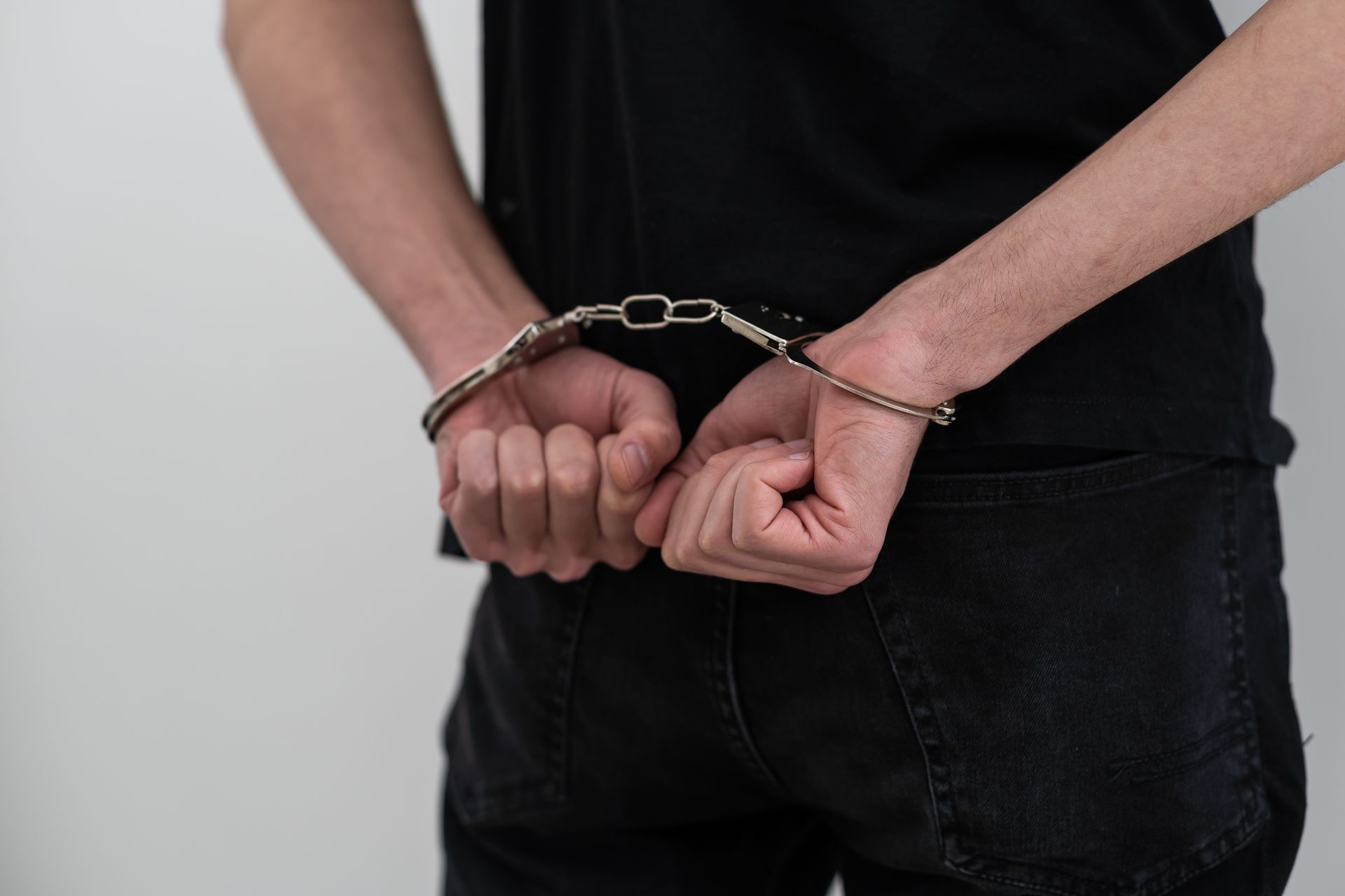 Person in black clothing, handcuffed behind the back, against a white background.