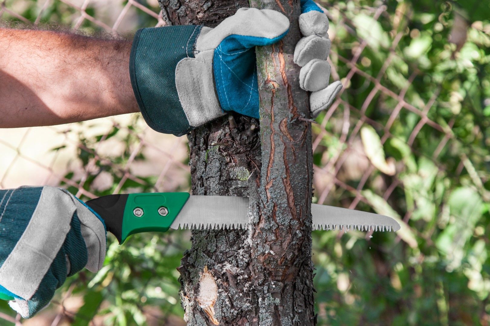 Gloved hands use a handsaw to cut through a tree trunk outdoors.