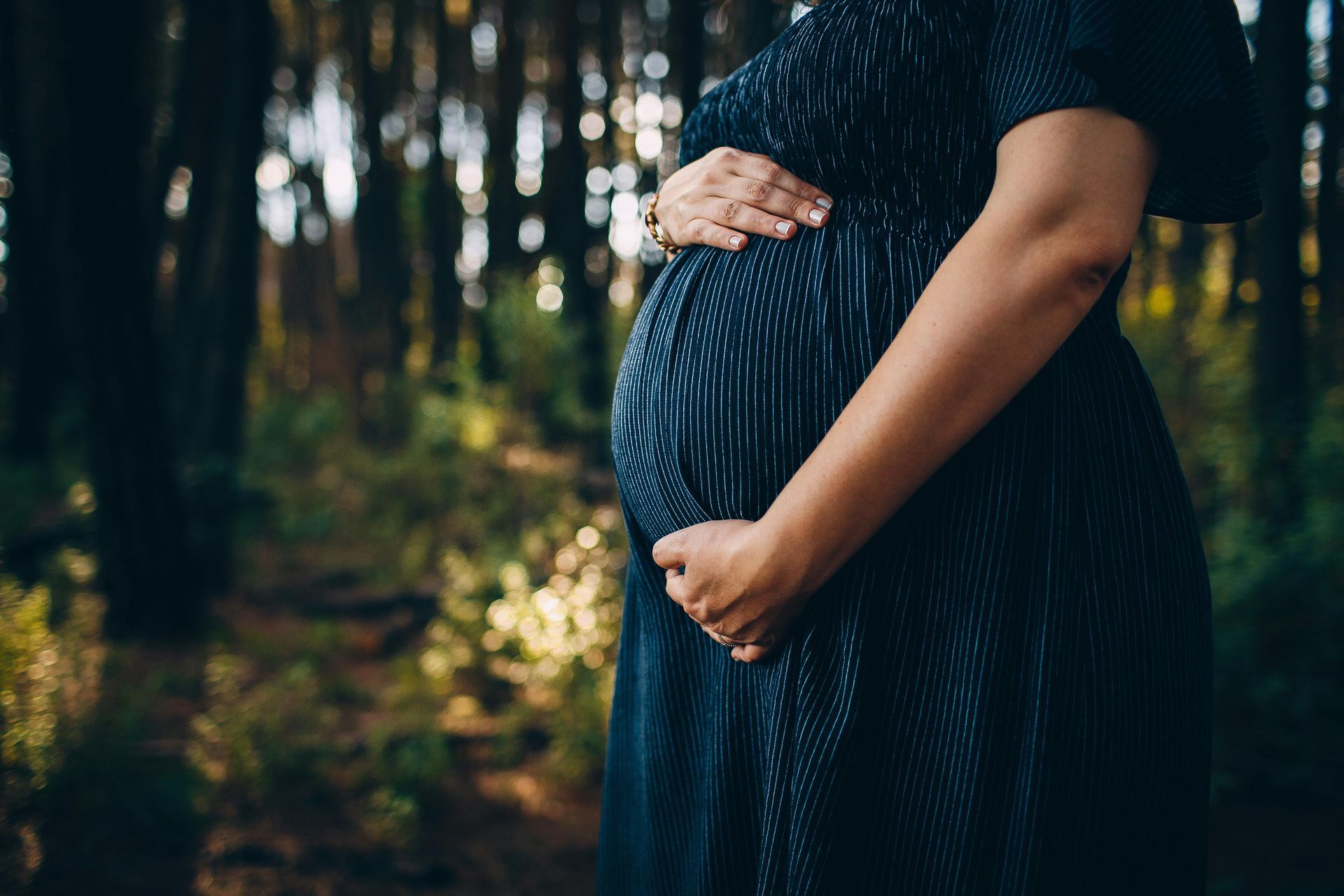 Pregnant woman in a dark blue dress, hands on belly, in a forest.