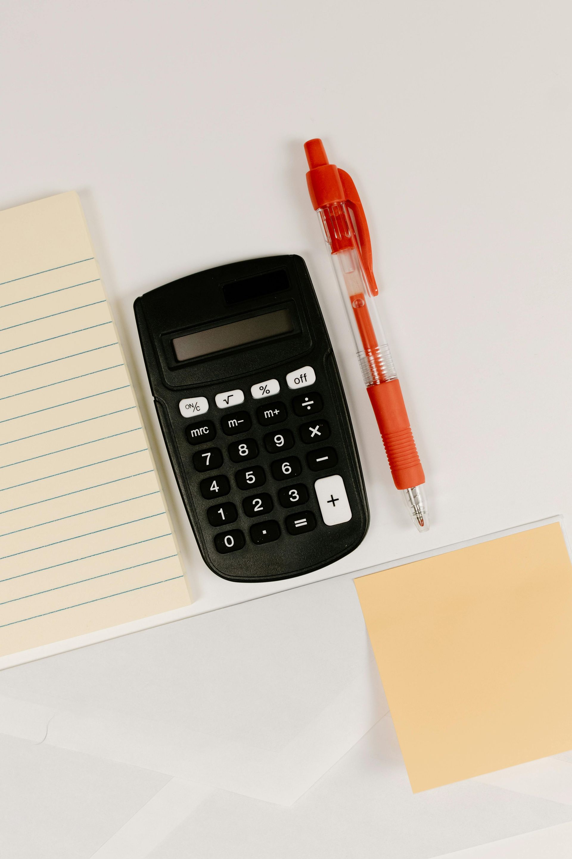 Black calculator, red pen, notepad, and sticky note on a white surface.