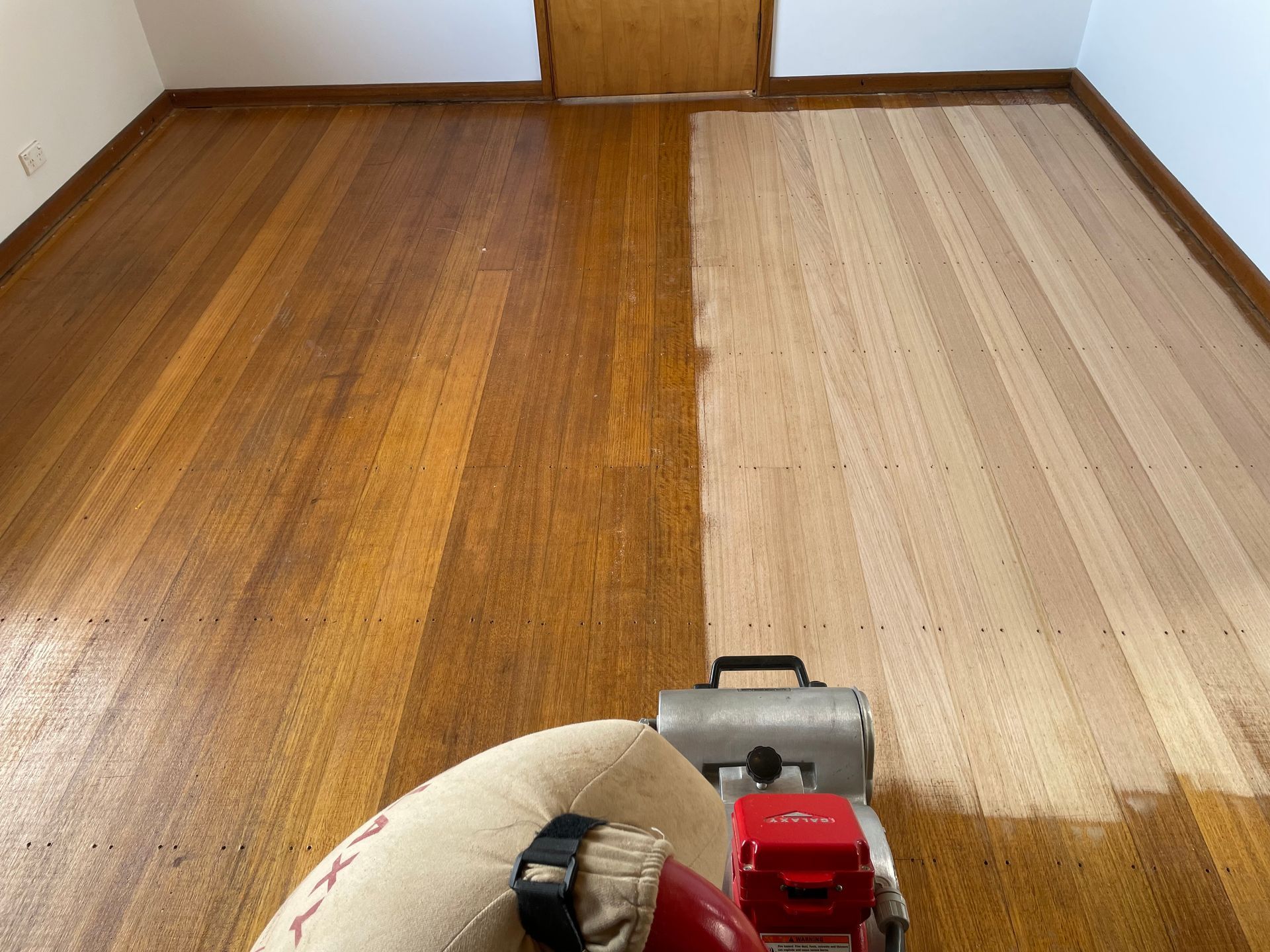 Person sanding a wooden floor, with a dark refinished section next to a lighter unfinished section. — Finer Floors In Cambridge, TAS