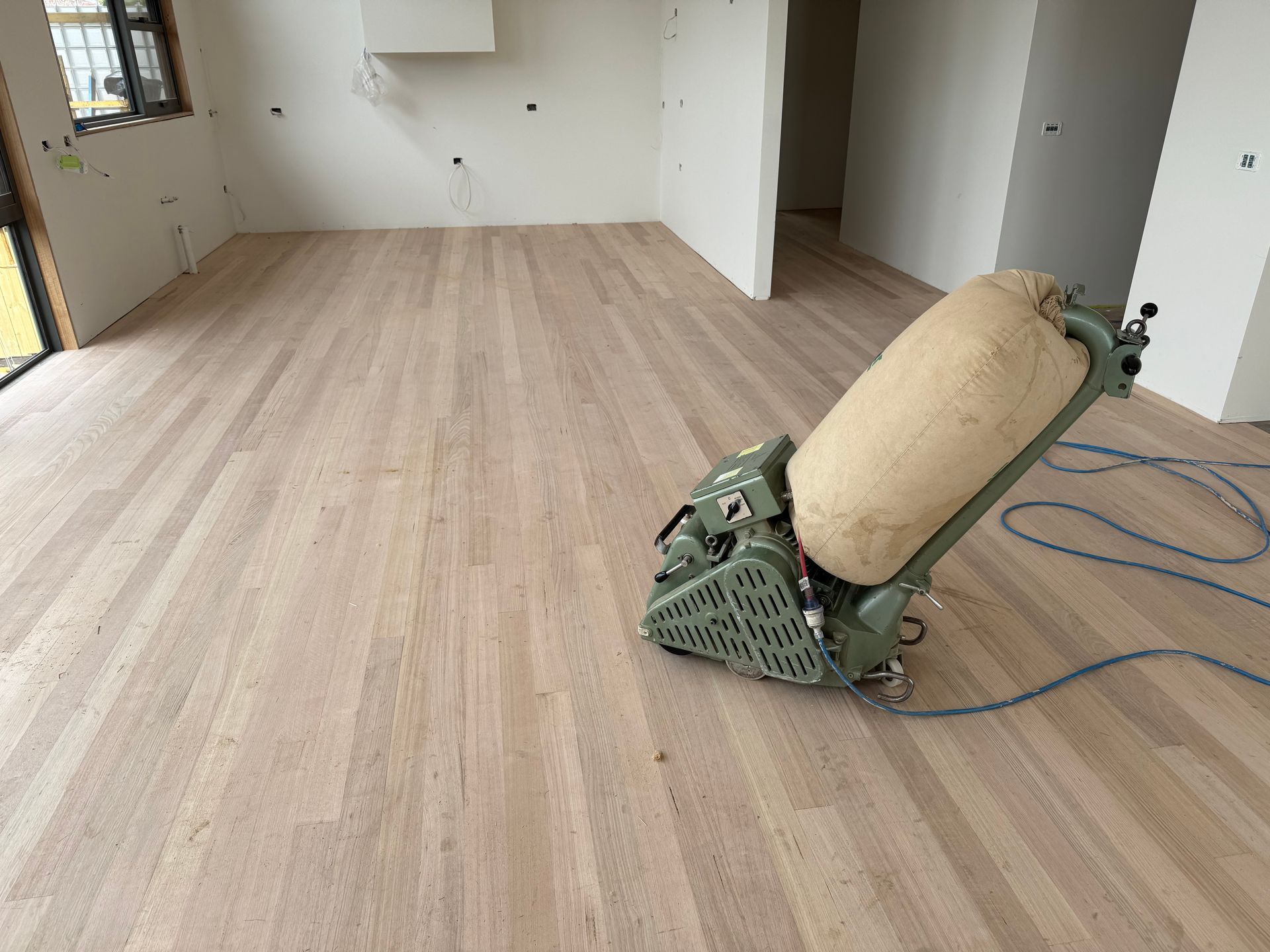 Empty room with light wood flooring being sanded by a floor sander and blue power cord. — Finer Floors In Cambridge, TAS
