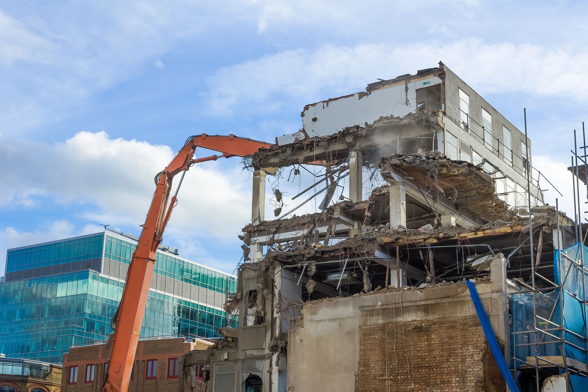 Escavatore che demolisce un edificio danneggiato accanto a un ufficio con facciata in vetro blu