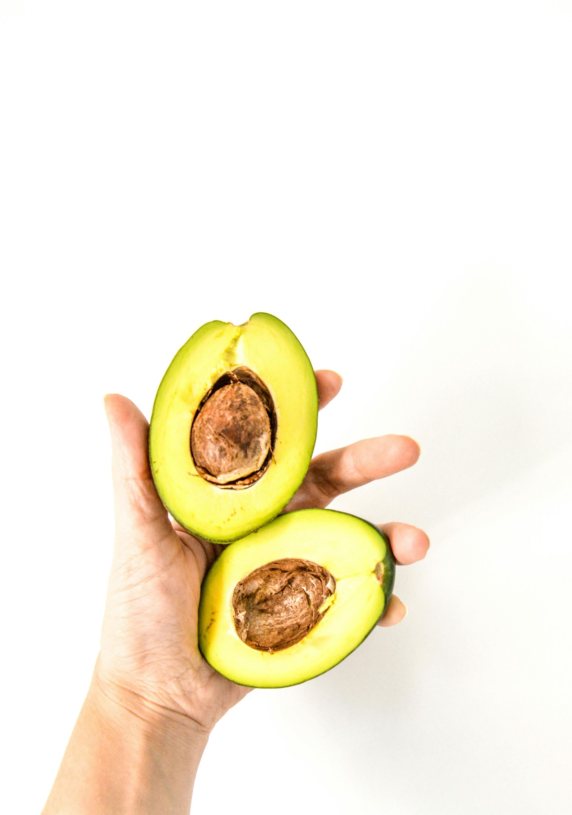 Hand holding a halved avocado, cut side up, showing the pit, against a white background.