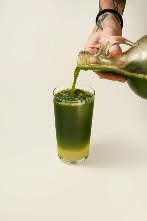 A person pouring green liquid from a glass pitcher into a glass with ice; light beige background.
