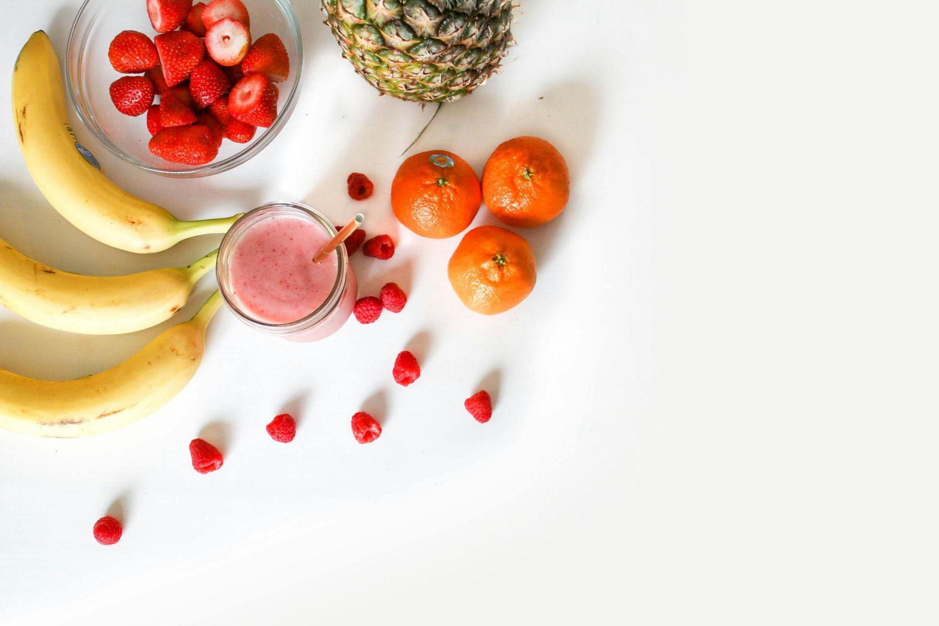 Bananas, strawberries, pineapple, oranges, smoothie, on white surface.