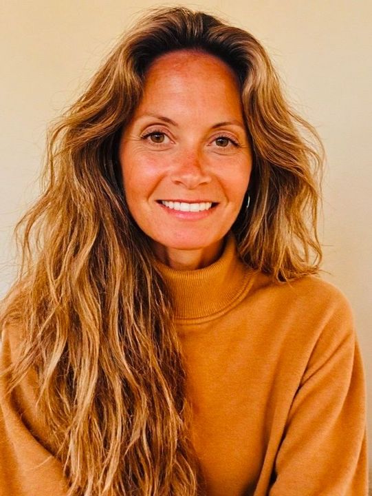 Smiling woman with long wavy hair wearing a tan sweater against a beige background