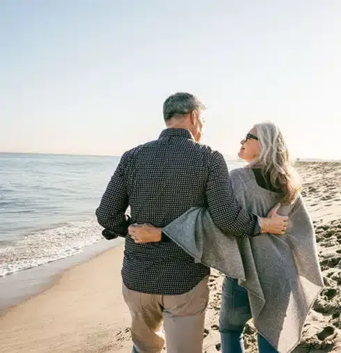 Couple walking arm in arm on a sandy beach at sunset, looking at the water