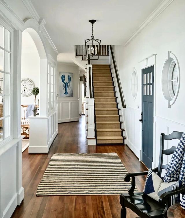 Bright hallway with staircase, striped rug, white walls, and black rocking chair.