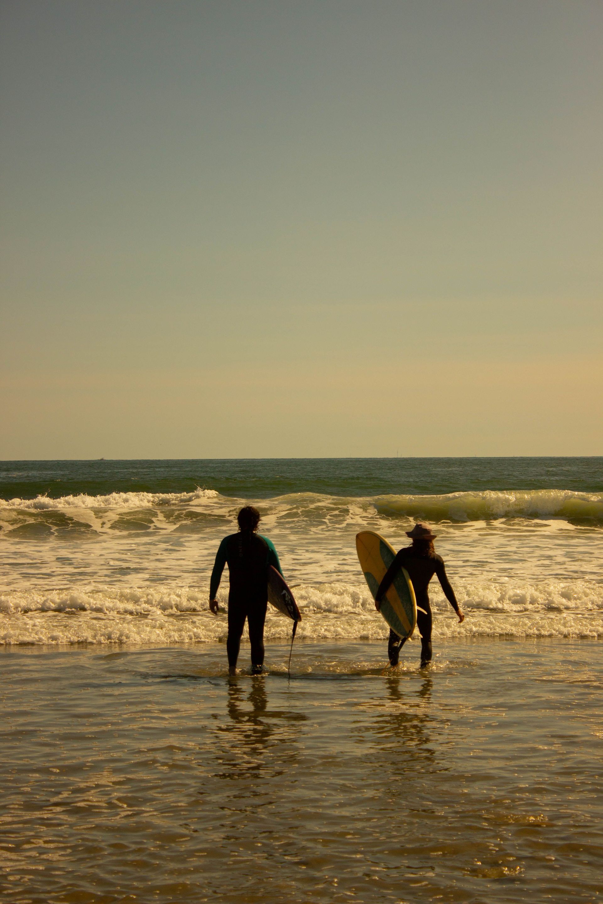 Two surfers walking into the ocean at sunset with boards, waves breaking ahead