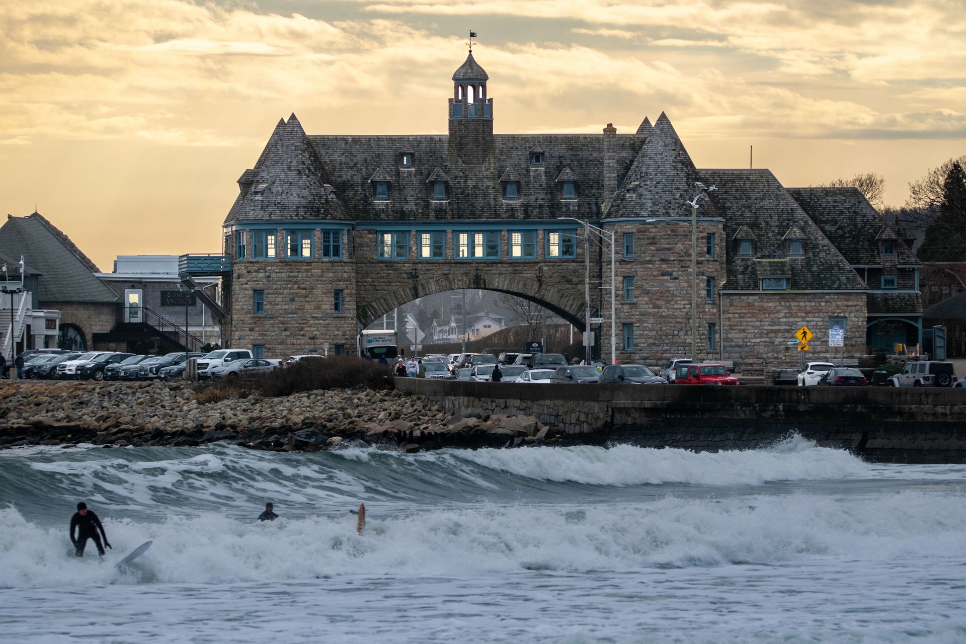 Stone seaside building with lit windows above rough waves and a lone surfer in the foreground at sunset