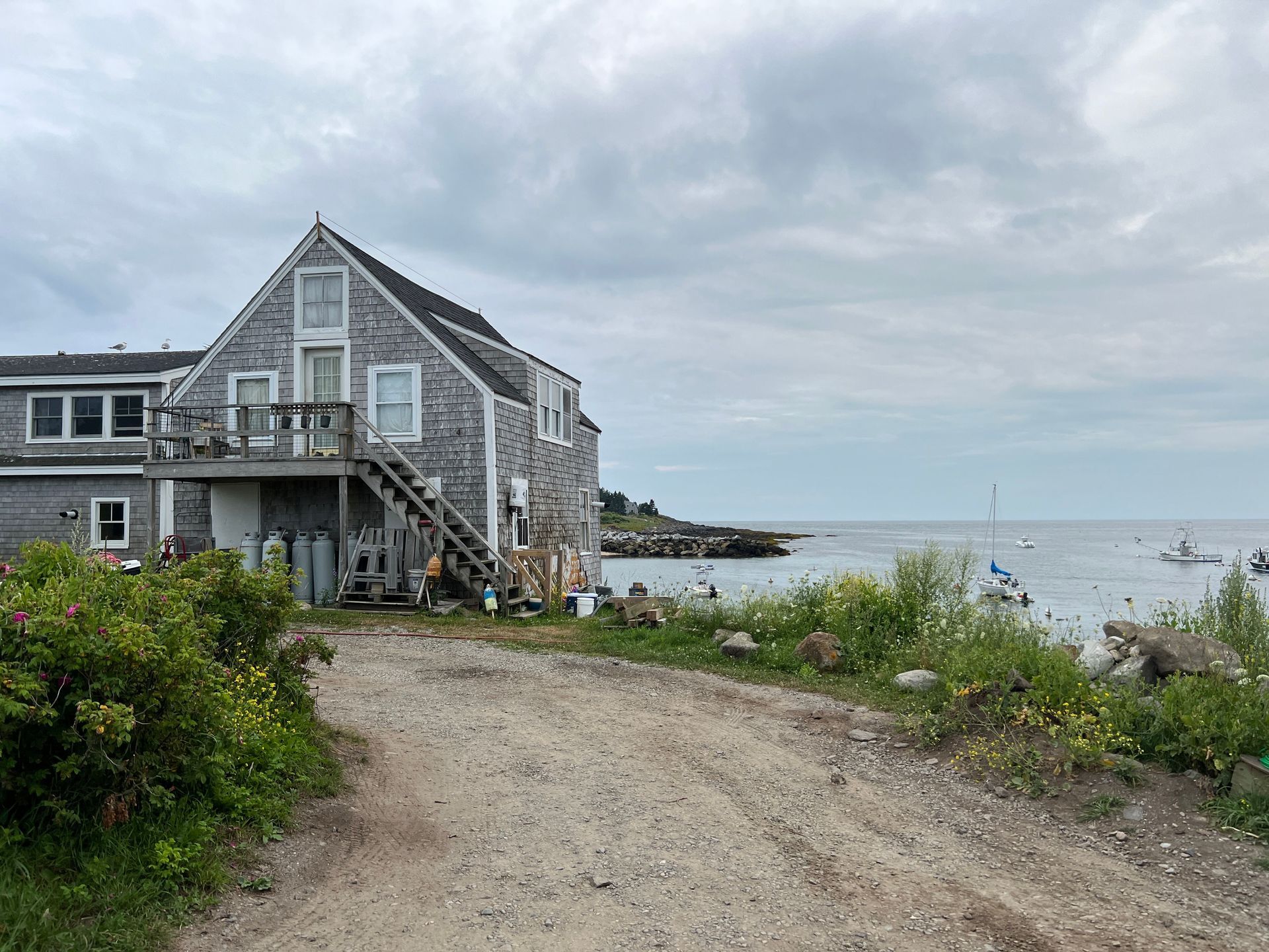 Coastal gray house beside a gravel path, with wildflowers and calm sea under a cloudy sky