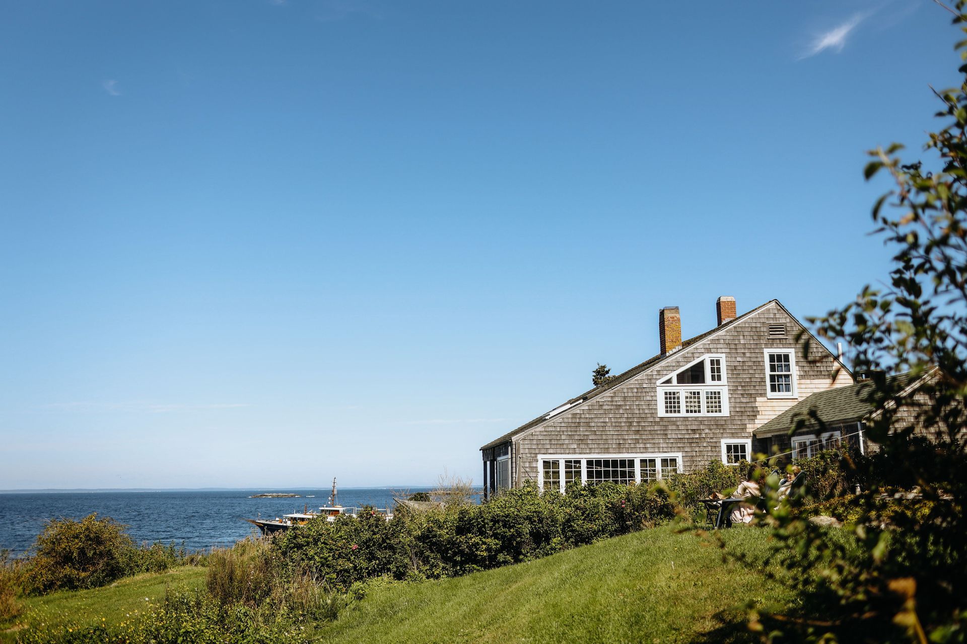 Coastal cottage beside the sea under a clear blue sky, with green shrubs in the foreground