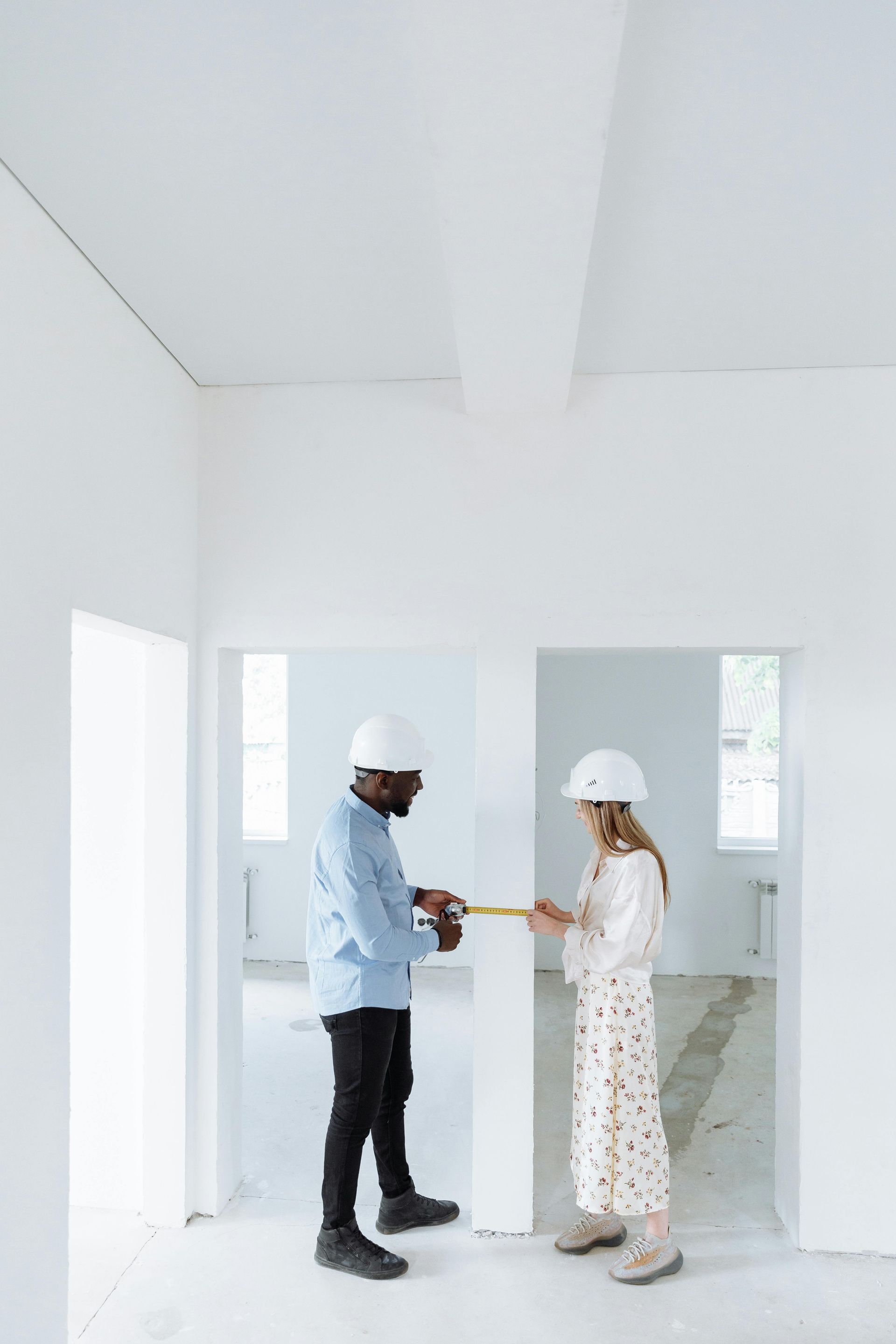 Two people in hard hats measuring a wall in a white room.