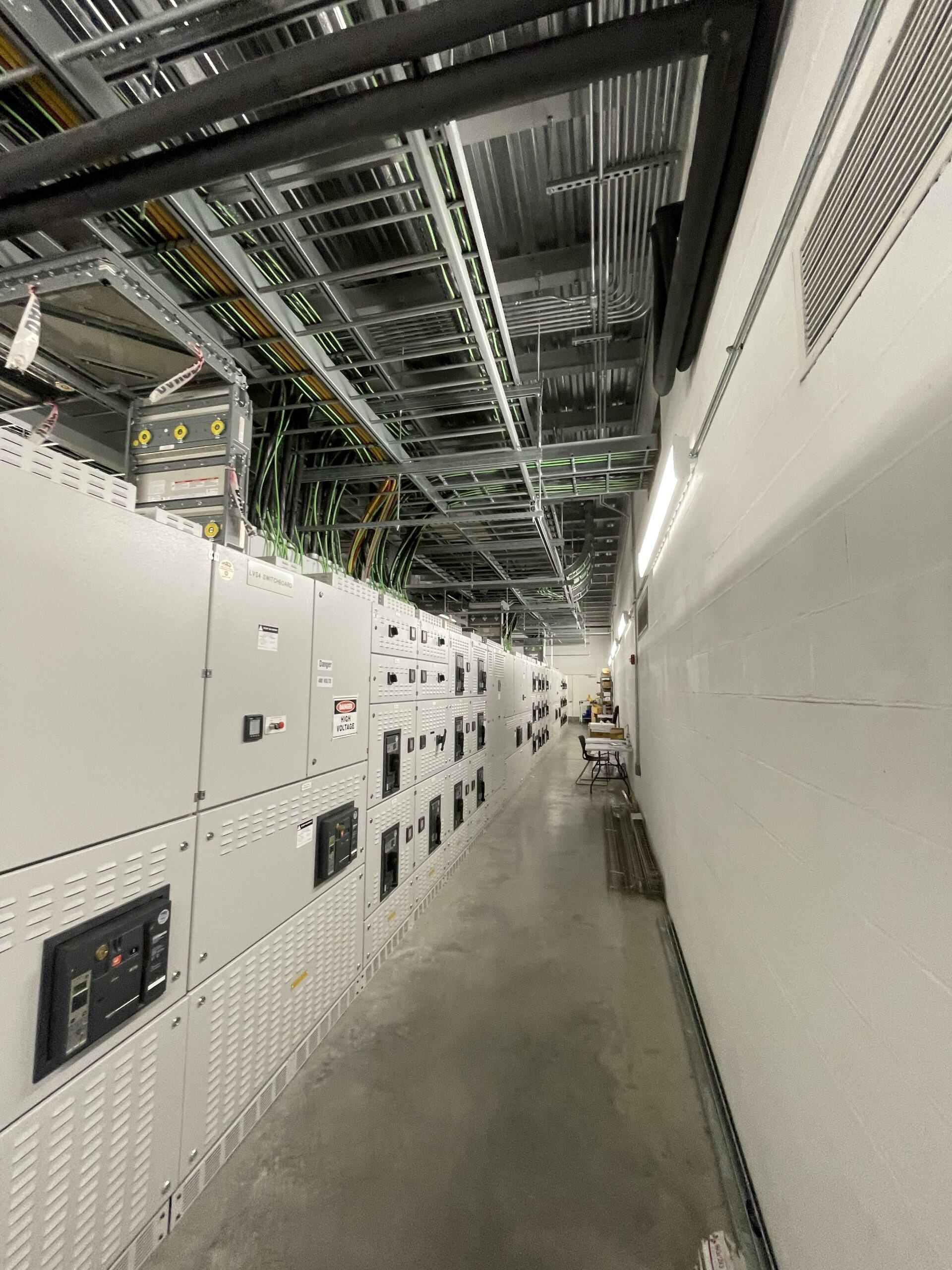 Narrow industrial corridor with white electrical cabinets, overhead cable trays, and a concrete floor