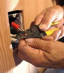 Hands using a wire stripper to work on electrical wiring inside a wooden wall.