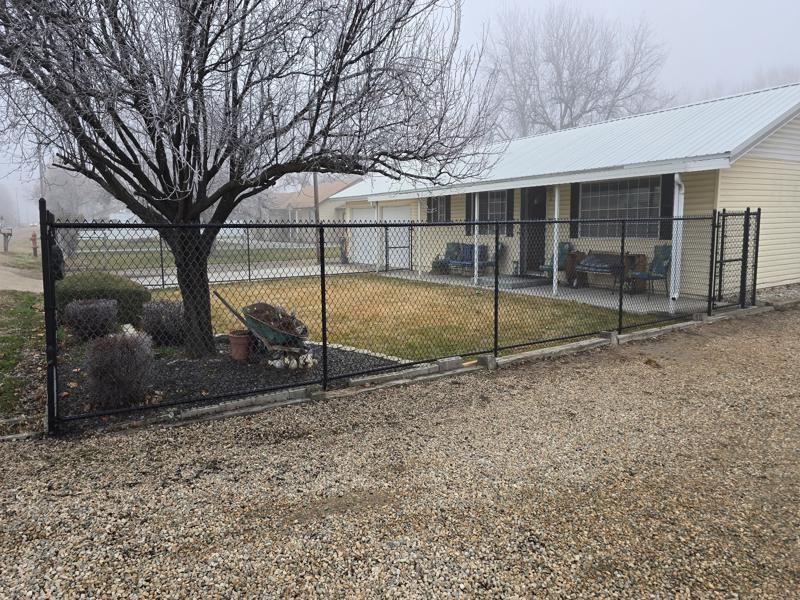 Chain-link fenced front yard with a leafless tree, gravel driveway, and beige house with porch