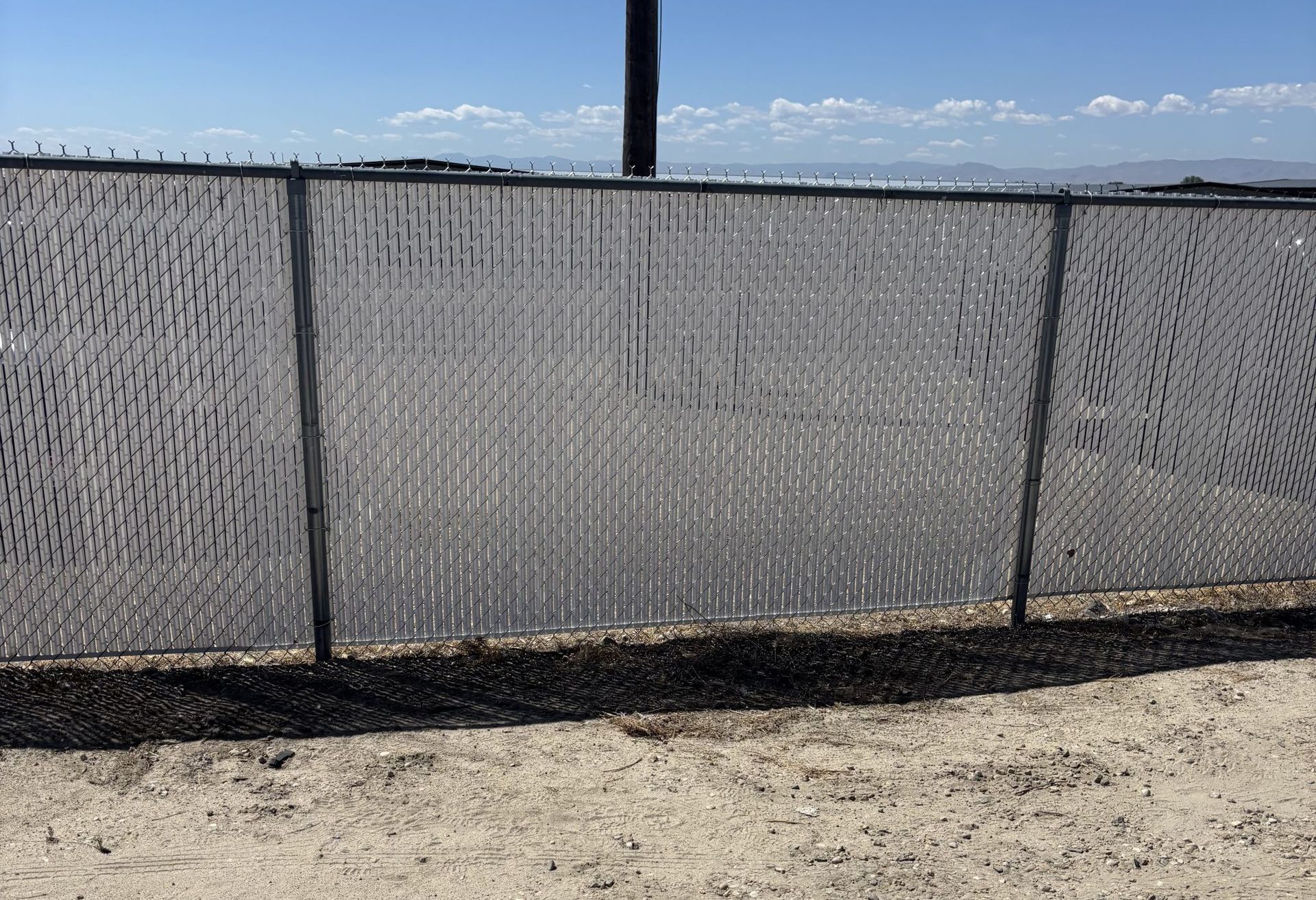 Chain-link fence with gray privacy mesh beside a dusty lot under a clear blue sky