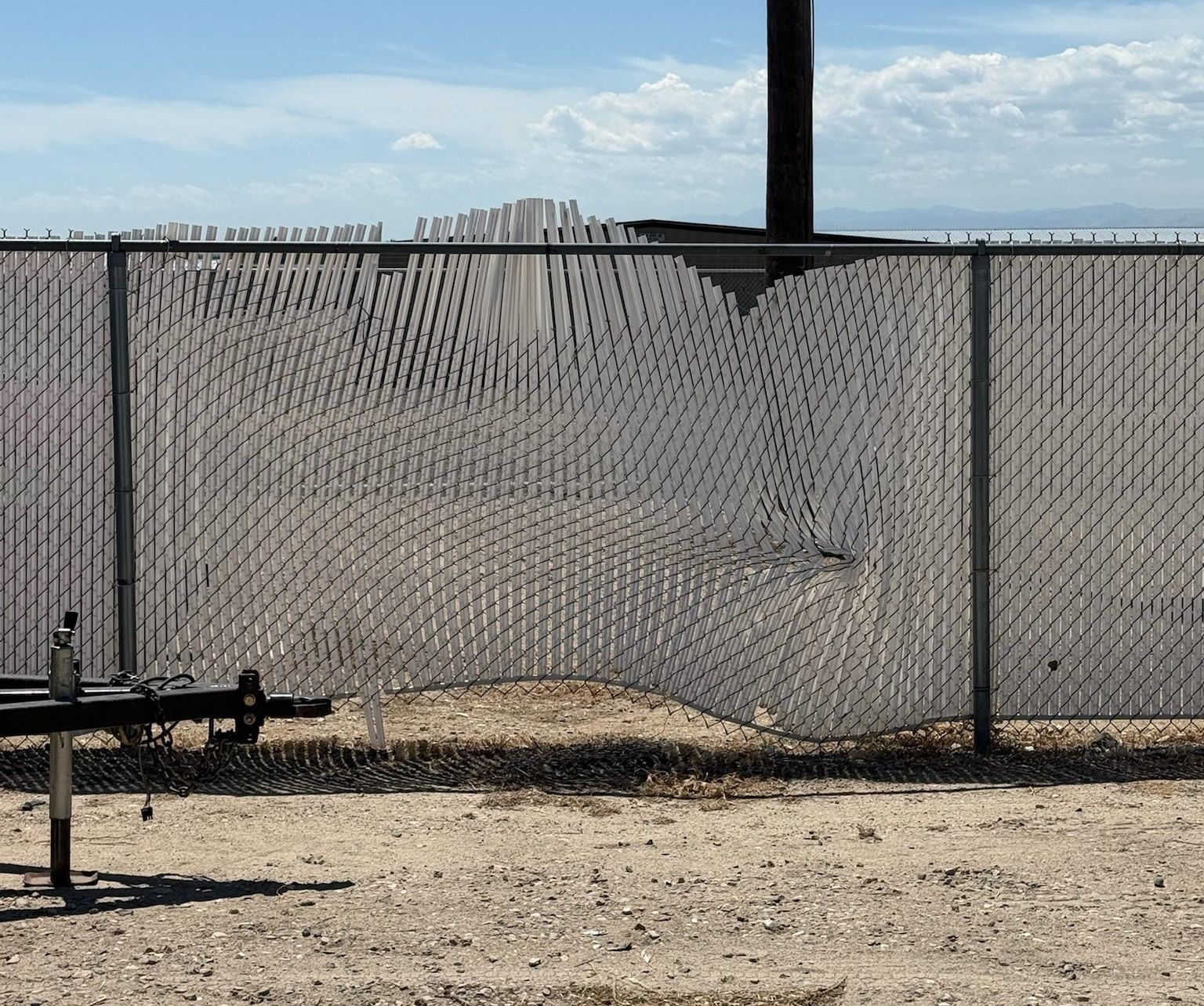 Chain-link fence with a large tear beside a dusty field under a bright sky.