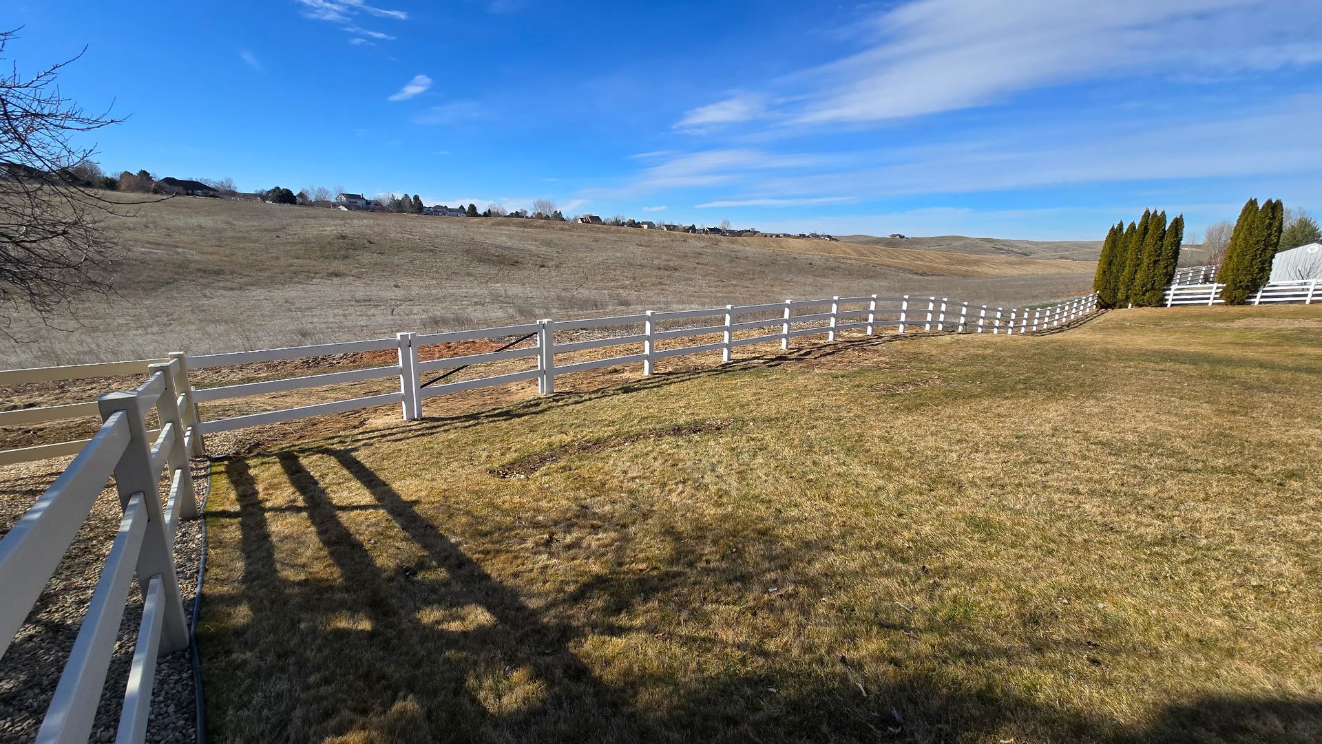 White fence lining a grassy hillside under a clear blue sky, with trees at right.