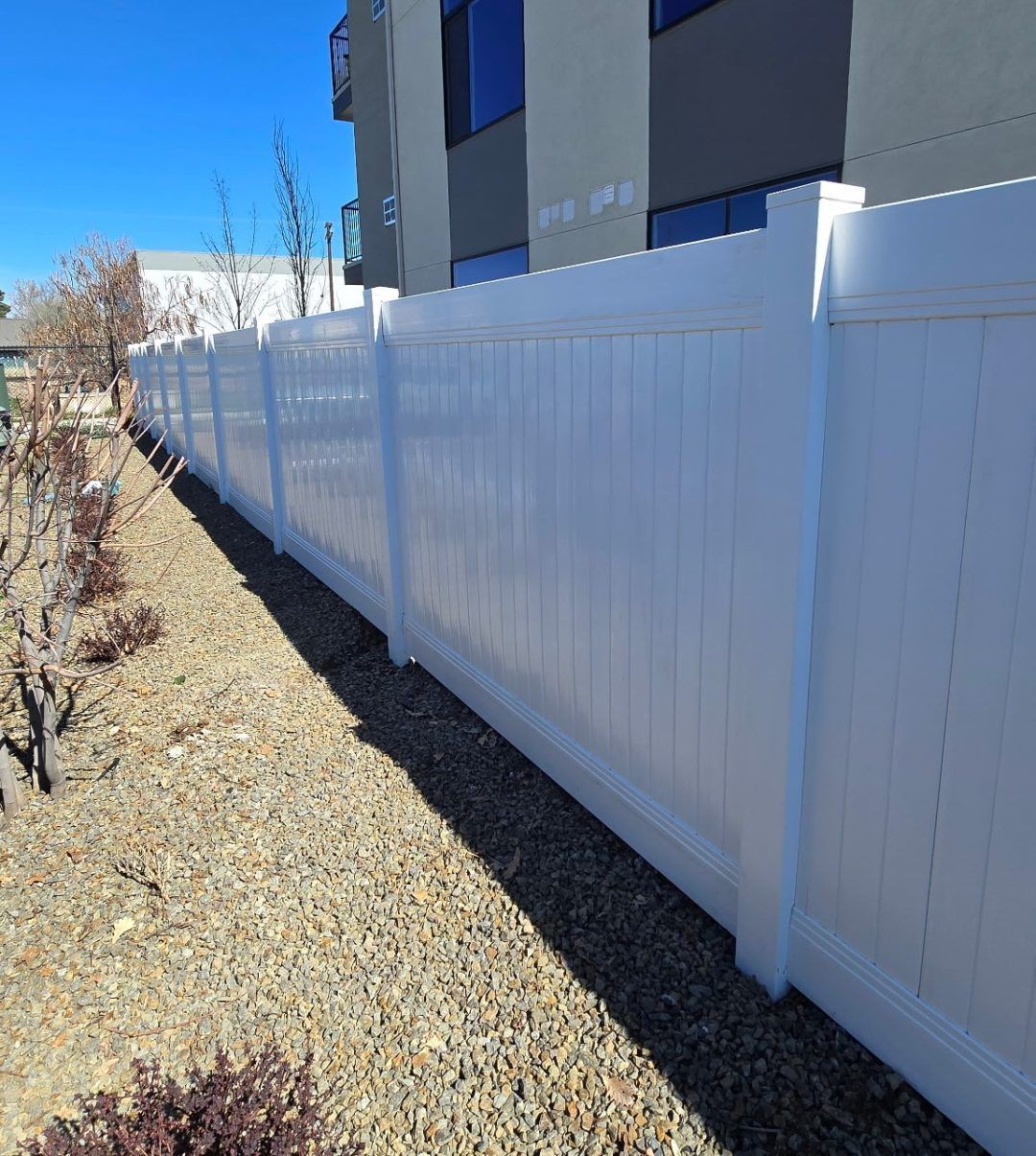 White vinyl fence beside a gravel path, with apartment buildings and shrubs in bright sunlight.