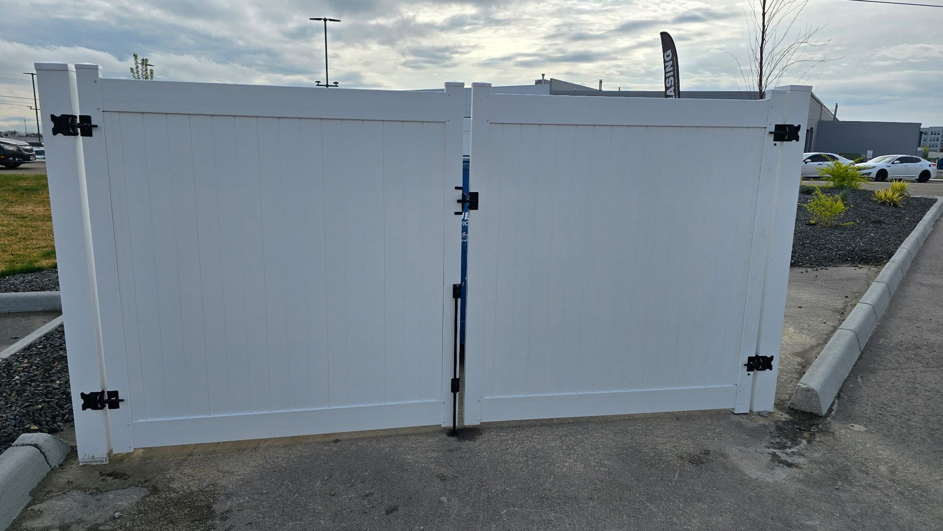 White double metal gate across a parking area, with black hinges and a gravel curb in front.
