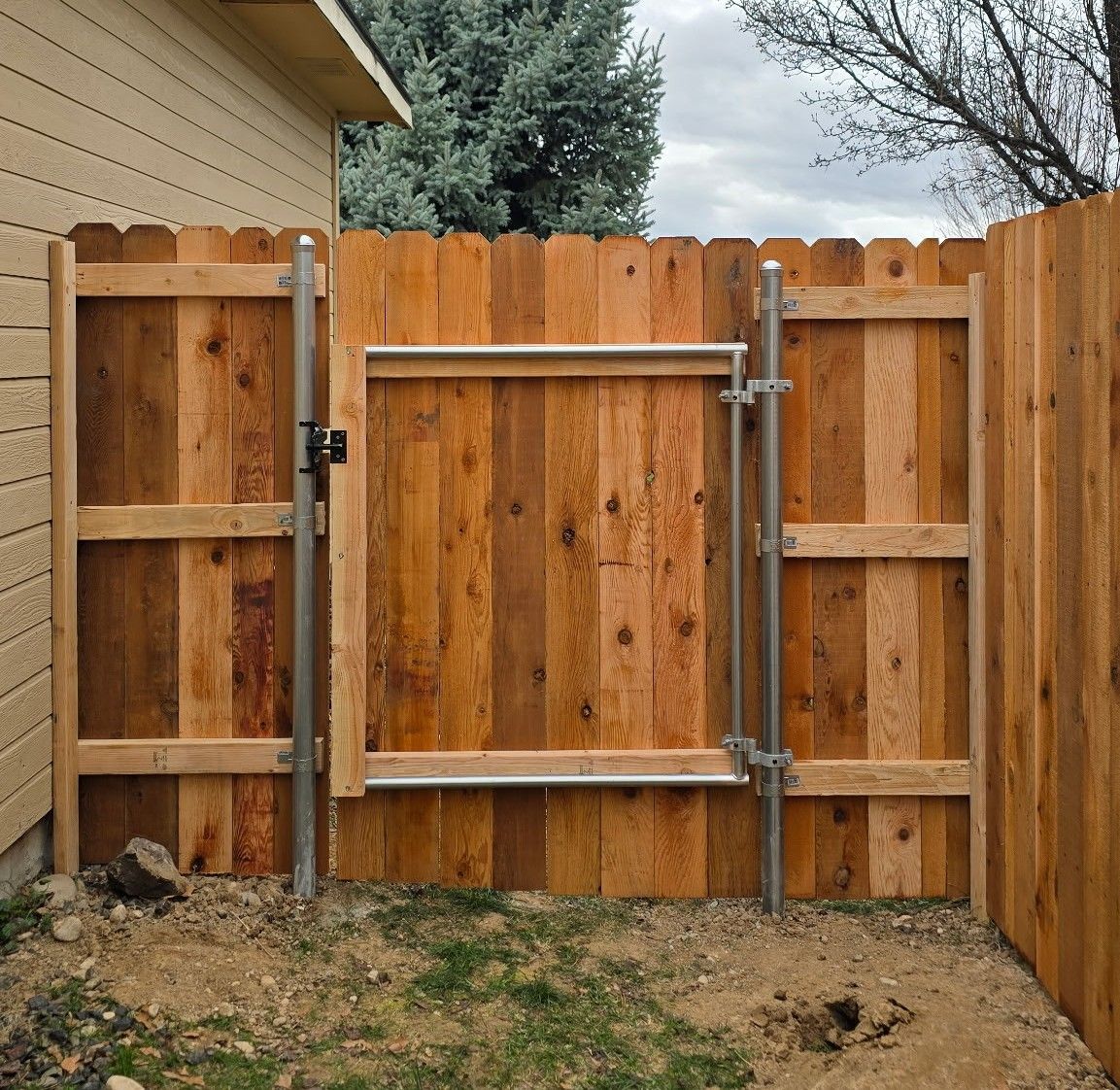 Wooden backyard fence with a metal gate and latch, viewed from a small dirt yard.