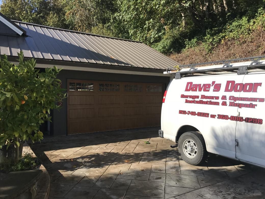 White service van parked beside a garage with a brown door and metal roof, under trees.