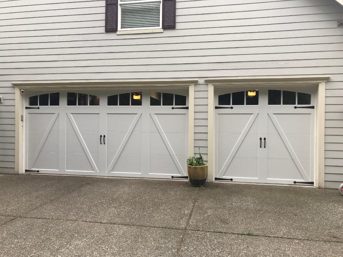 Two white garage doors with windows on a gray house exterior, with a potted plant between them