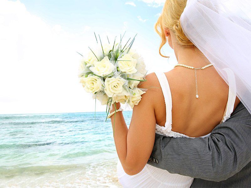 A bride and groom are hugging on the beach and the bride is holding a bouquet of white roses
