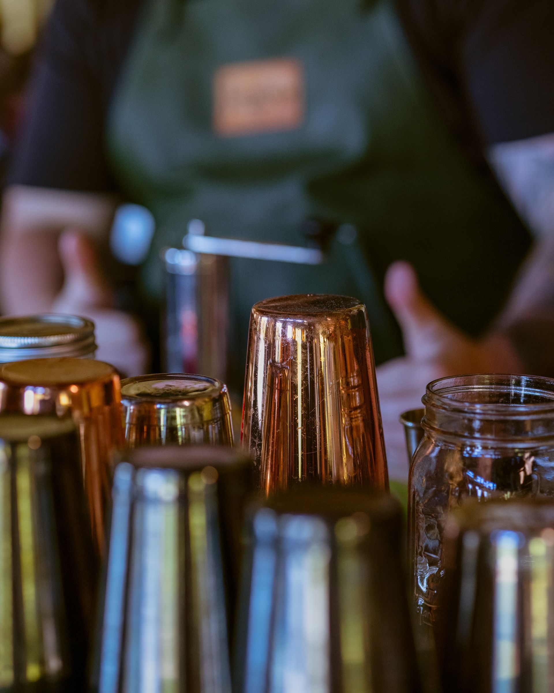 Bar scene with copper and metal shakers in focus, bartender blurred in background, wearing a green apron.