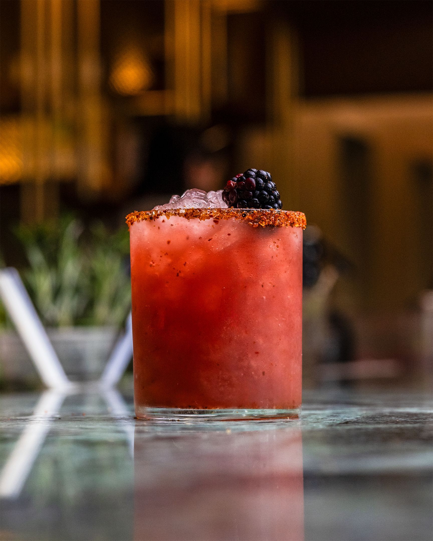 Red cocktail in a glass with a sugared rim, garnished with a blackberry, on a reflective surface.