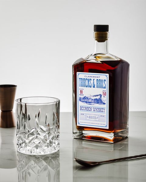 Bottle of liquor, a rocks glass, jigger, and spoon on a marble surface against a white background.