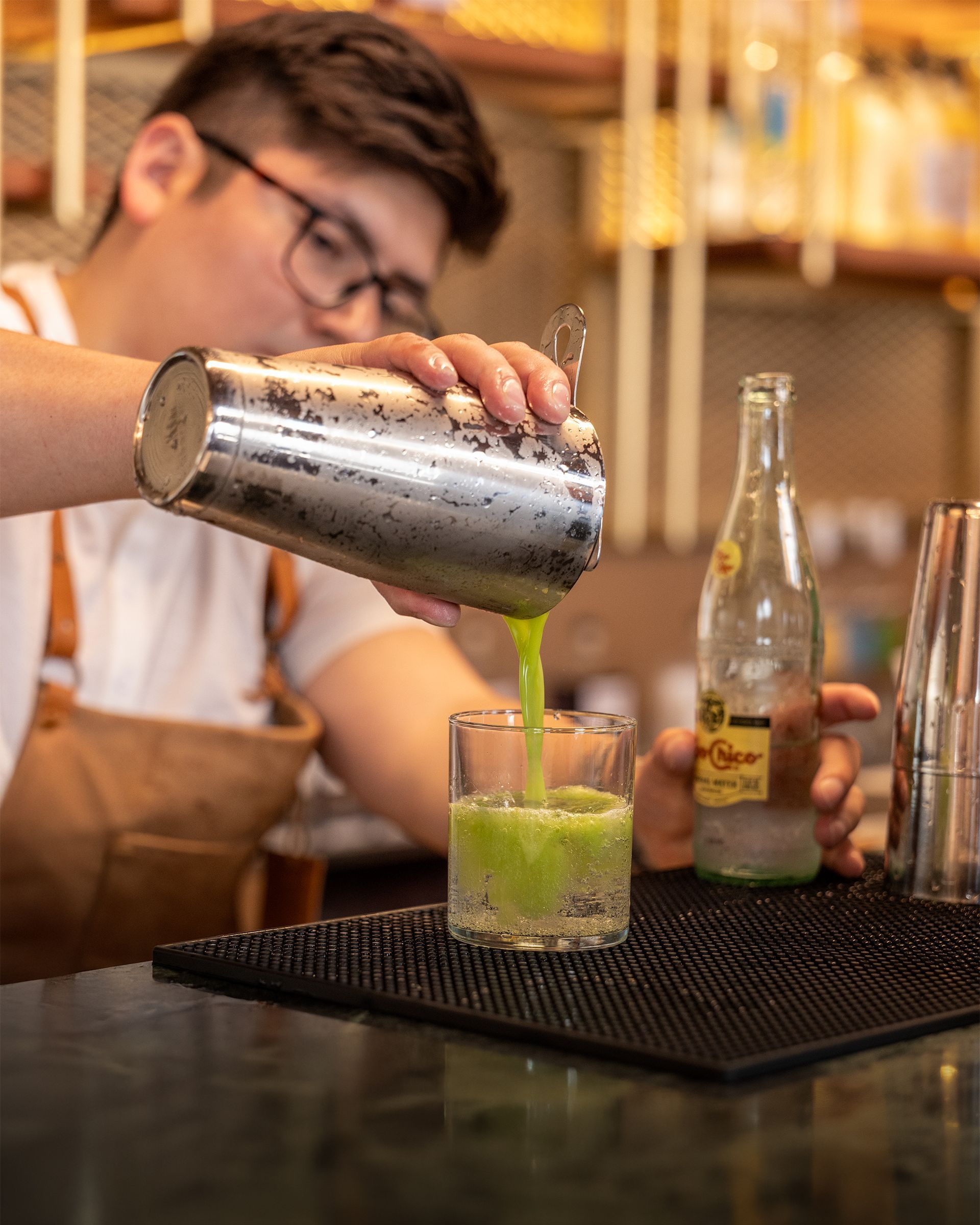 Bartender pouring green cocktail from shaker into glass. Bottle and other shaker nearby.