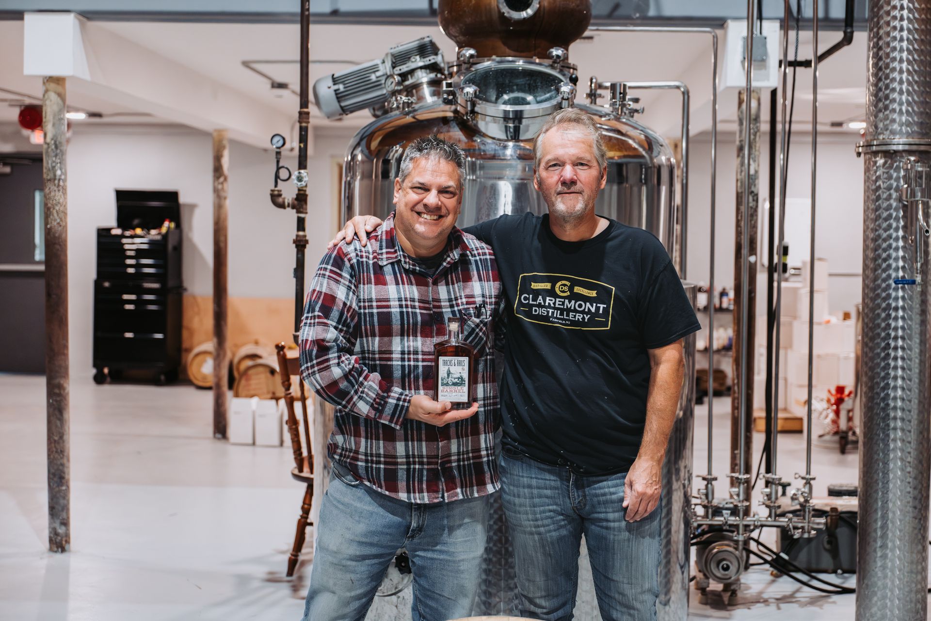 Two men pose with a bottle in a distillery, near a large still. One holds the bottle.