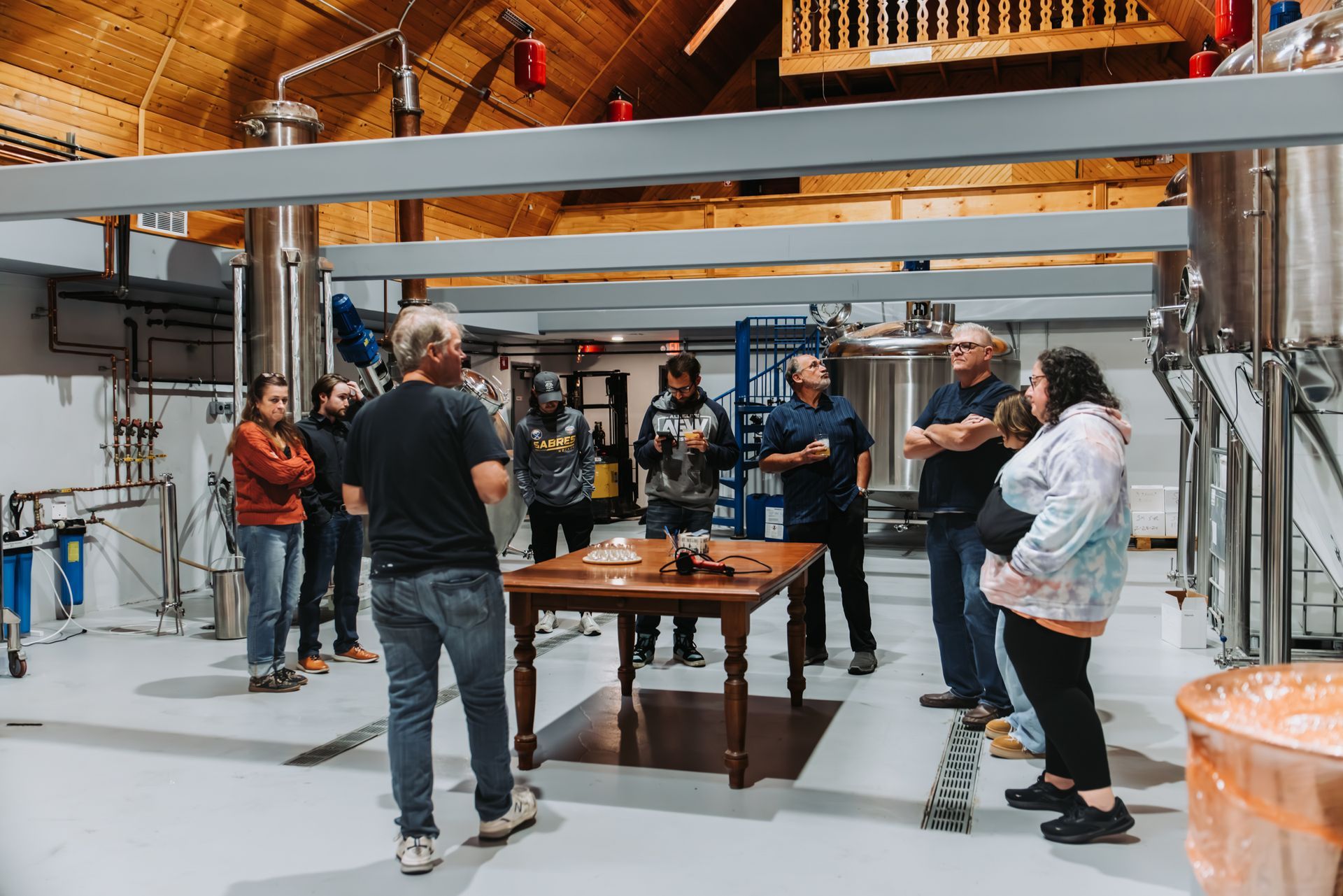 Group of people in a brewery, listening to a presentation at a table. Silver tanks and pipes visible.