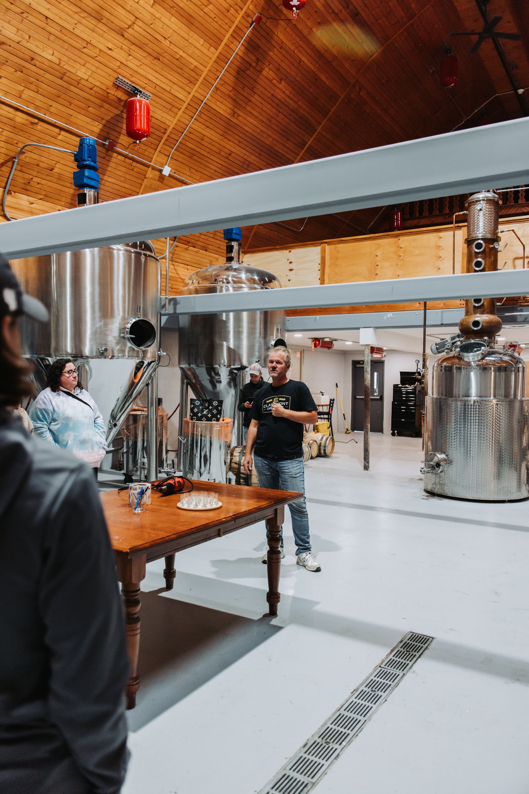 Tour guide in distillery, pointing, with stainless steel equipment and wood ceiling.