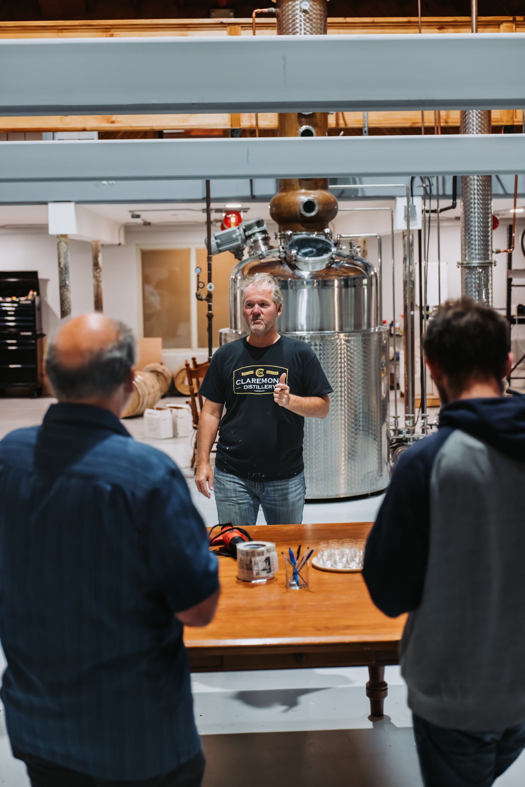 Man speaking to two others at a distillery, stainless steel equipment in the background.