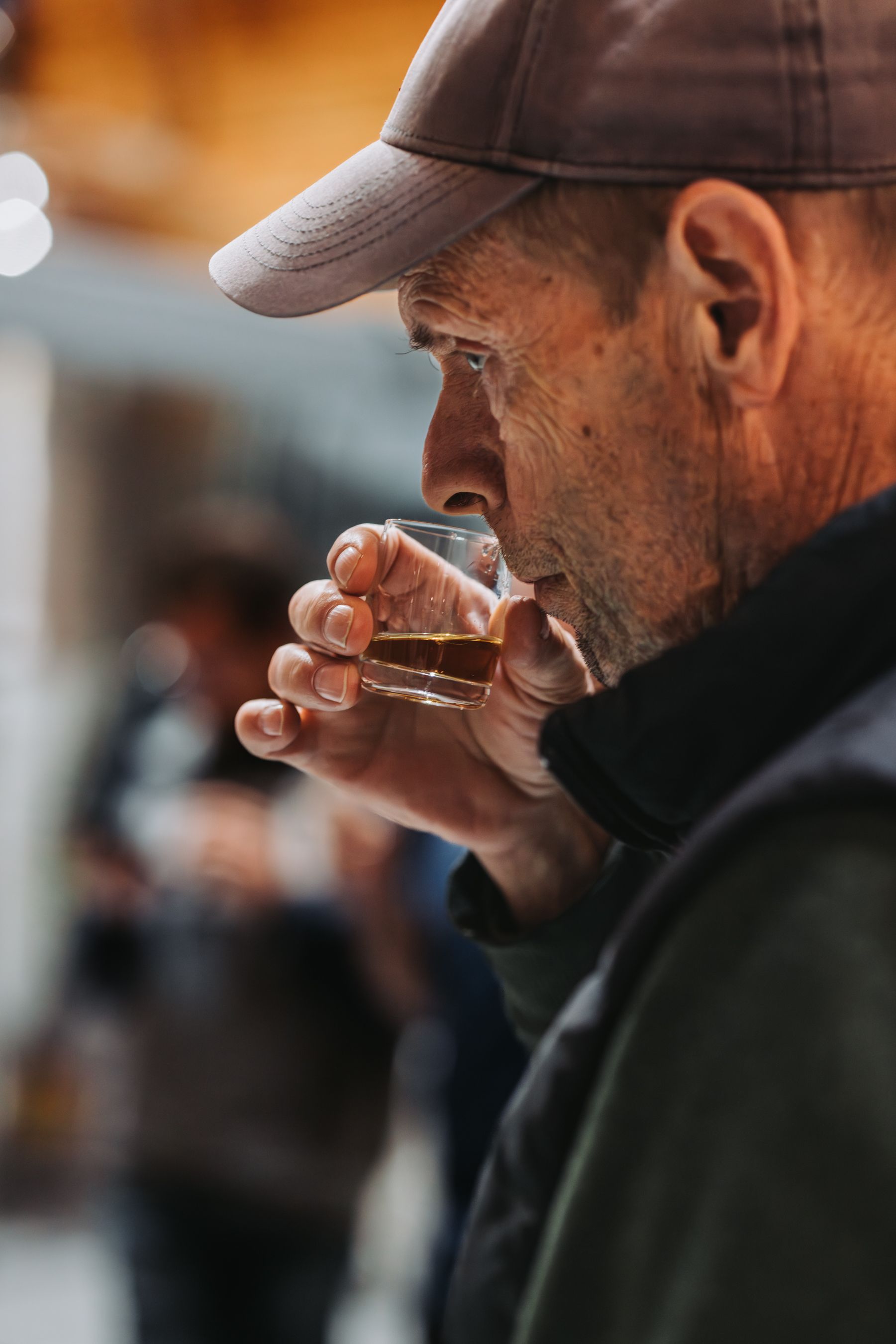 Man smelling a glass of amber liquid, wearing a cap, indoors.