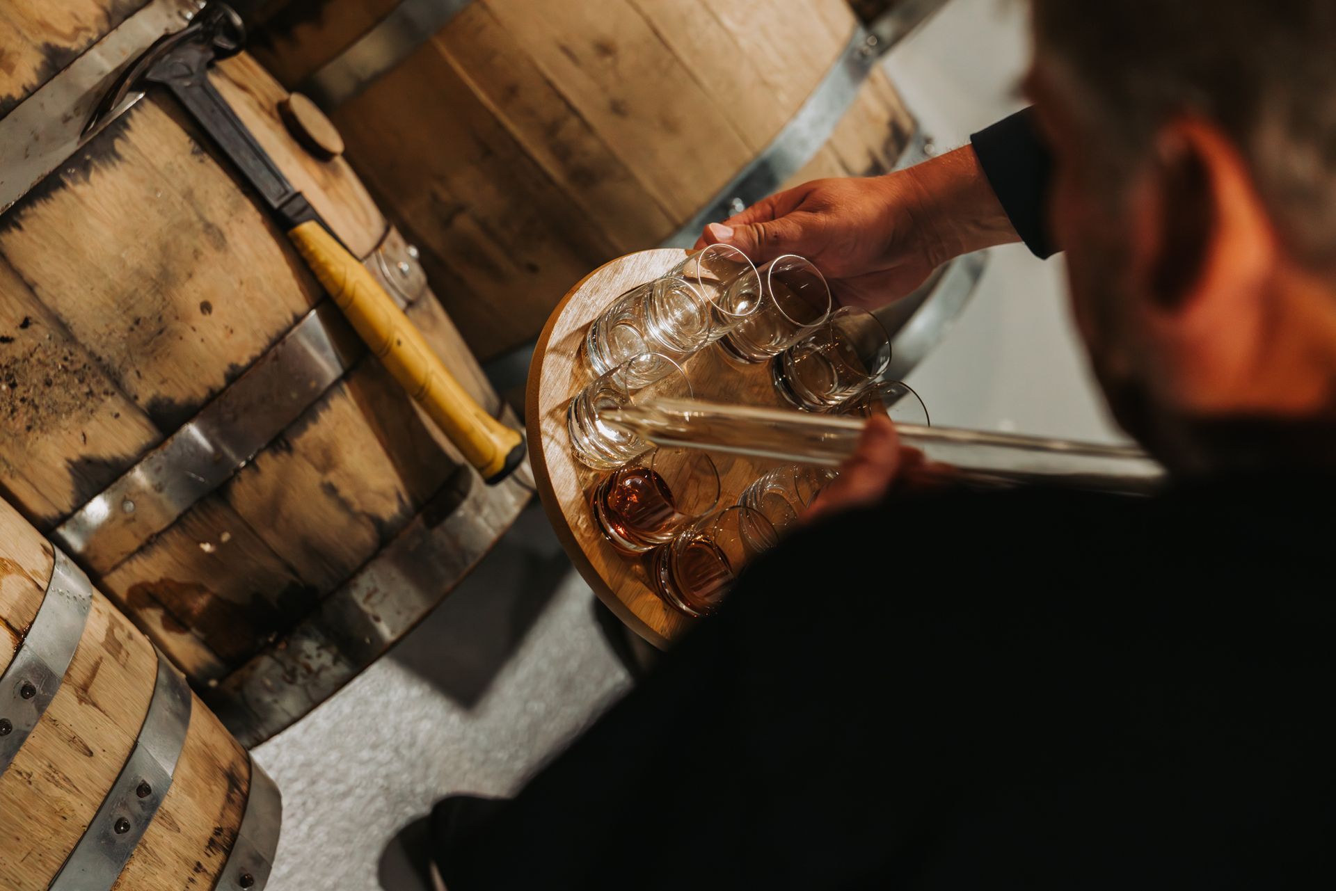 Person pouring amber liquid into glasses, beside whiskey barrels in a dimly lit room.