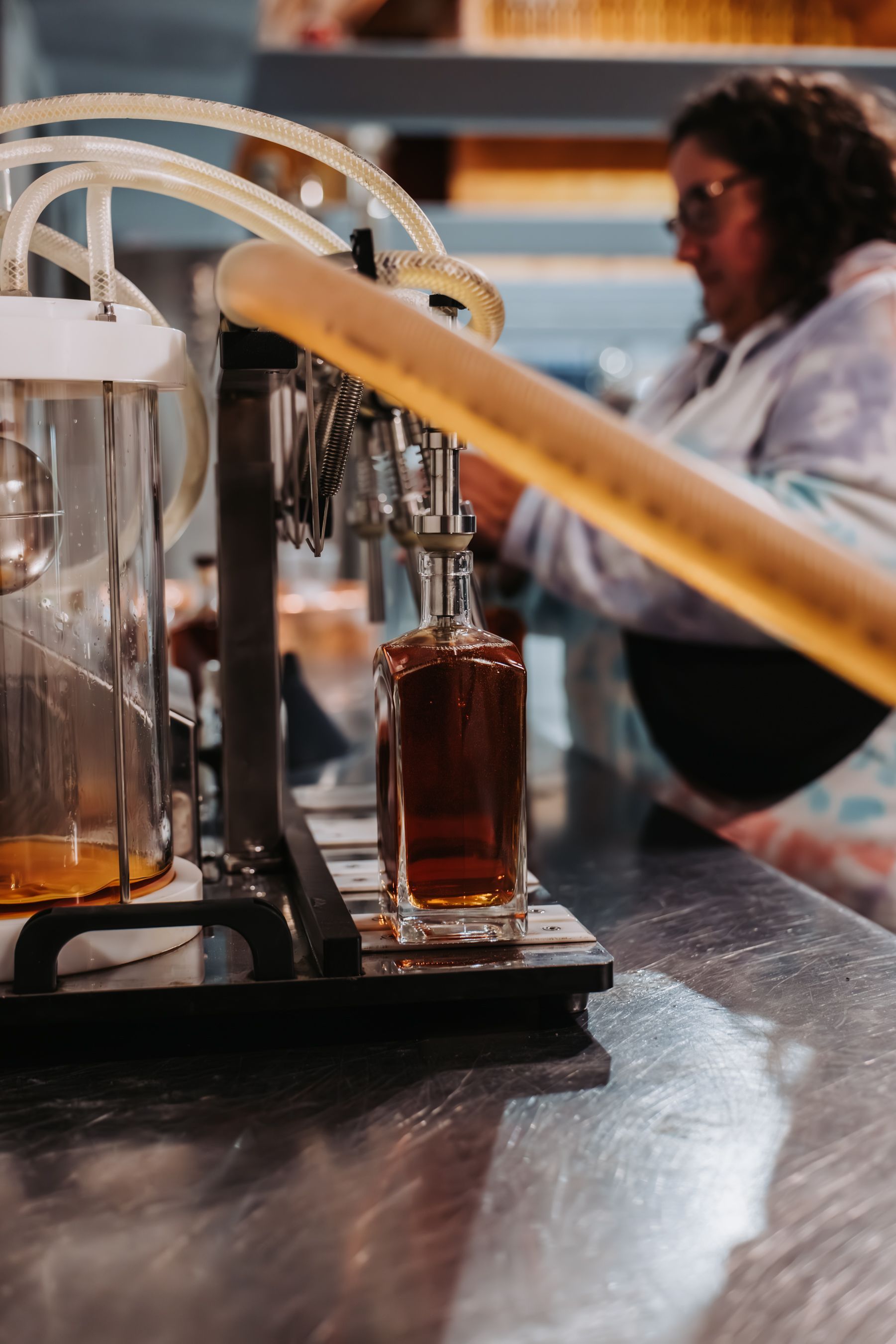 A bottle being filled with amber liquid from a machine, with a person in the background.