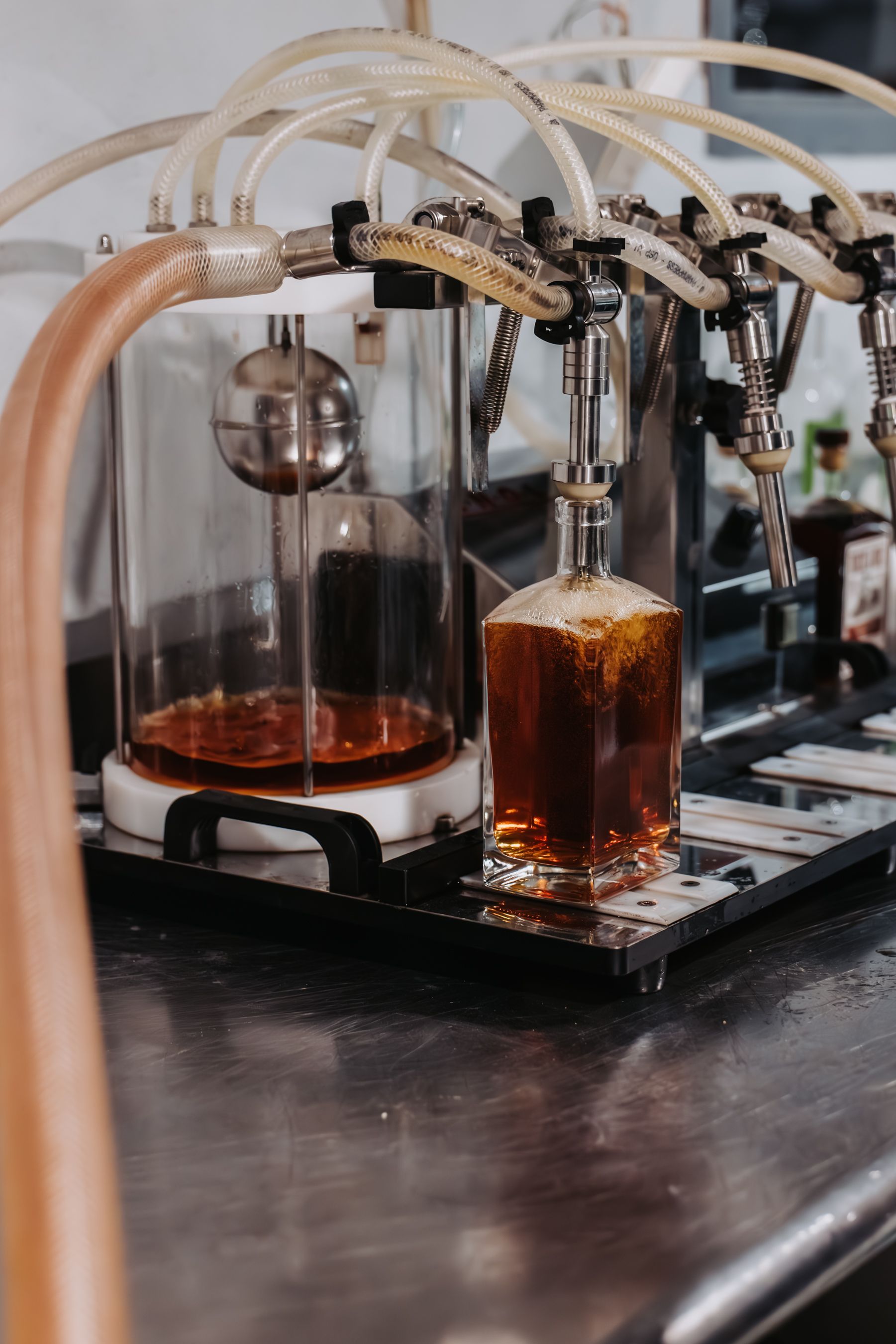 Automated pouring system filling a glass bottle with amber liquid. Clear tubes, steel fixtures, and a modern setting.