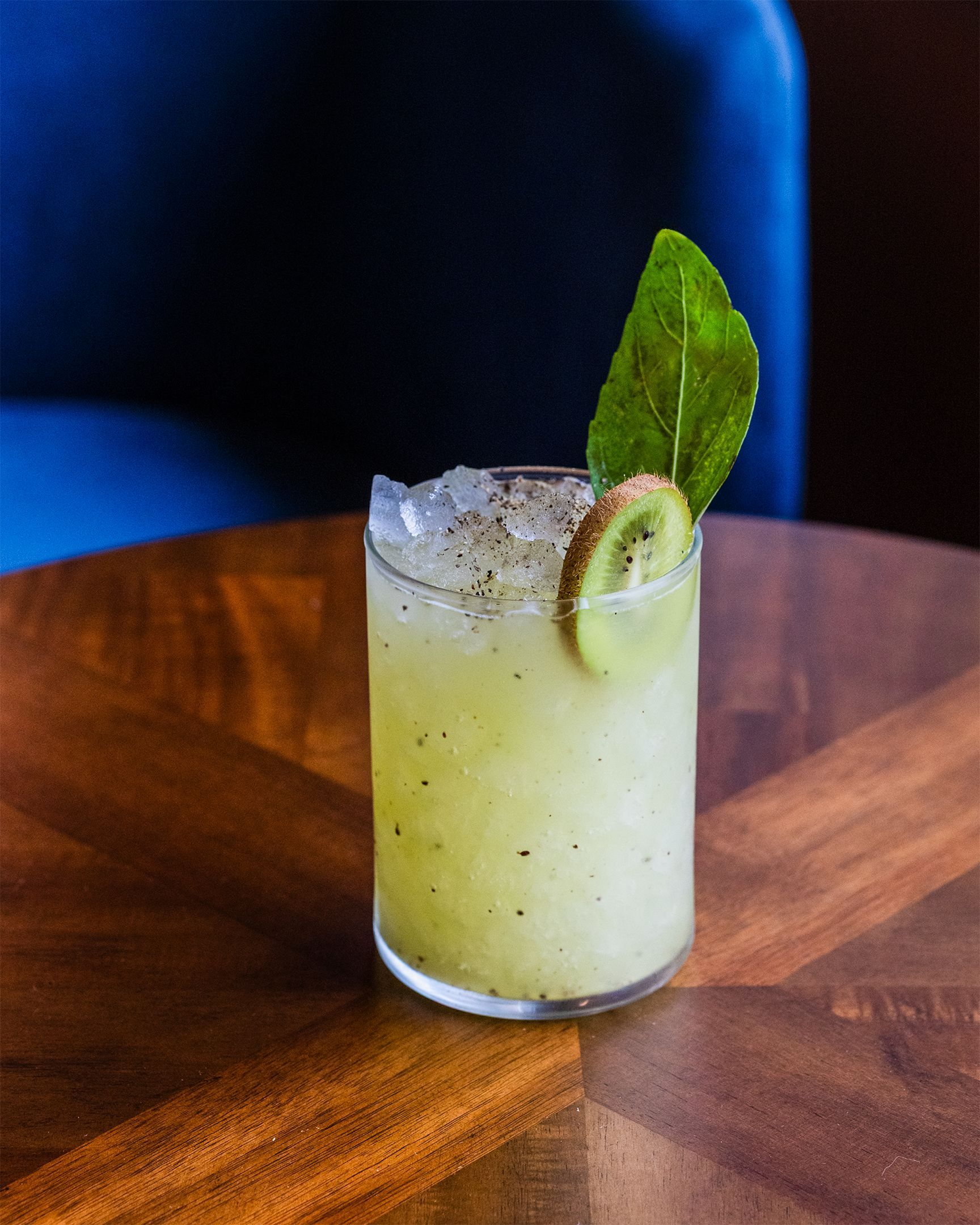 Kiwi cocktail in a clear glass, garnished with a kiwi slice and a green leaf, on a wooden table.