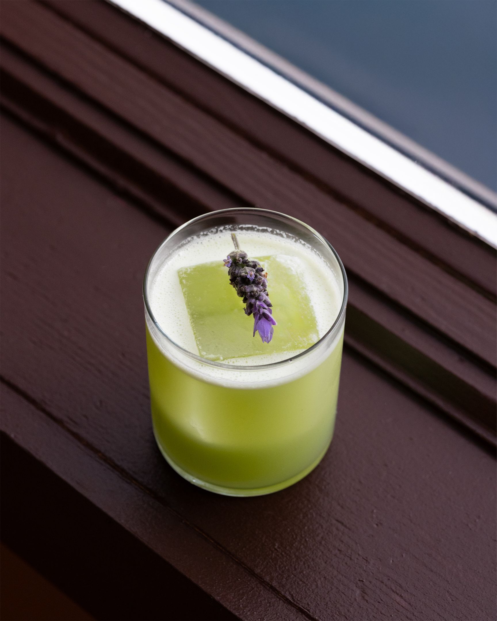 Green cocktail in a rocks glass, garnished with lavender, on a dark wood surface.