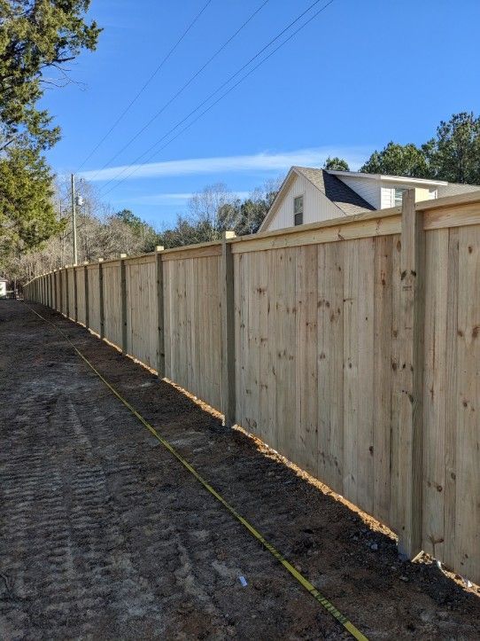 A newly installed wooden privacy fence runs along a dirt path under a clear blue sky.